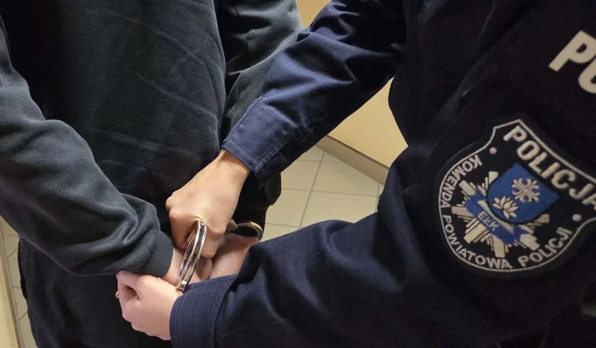 Funkcjonariusz policji z komendy powiatowej w Ełku zakłada kajdanki na ręce osoby ubranej w ciemny dres.