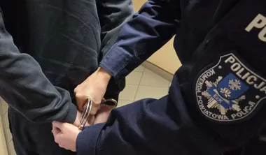 Funkcjonariusz policji z komendy powiatowej w Ełku zakłada kajdanki na ręce osoby ubranej w ciemny dres.
