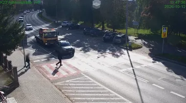 Piesi przechodzą przez przejście dla pieszych na skrzyżowaniu, gdzie samochód osobowy i pomoc drogowa czekają przed pasami, a inne pojazdy jadą w tle.